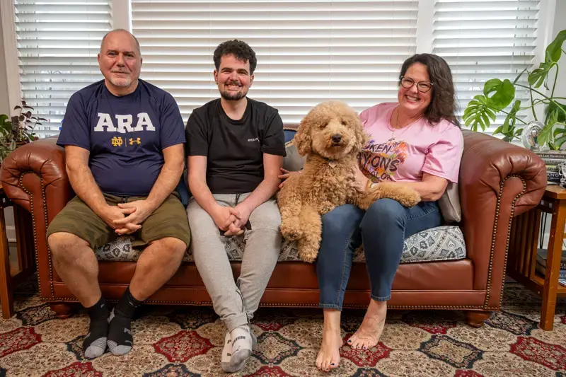 Harry, Alec and Gail Koujaian sit on a brown leather couch with their golden doodle dog.