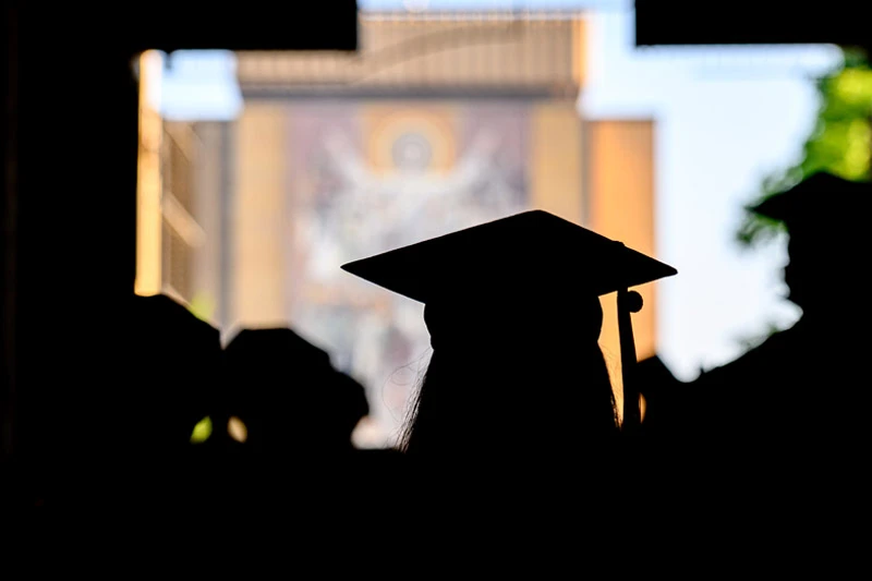 Silhouette of a graduating student wearing a mortarboard, with the Basilica of the Sacred Heart at Notre Dame blurred in the background.