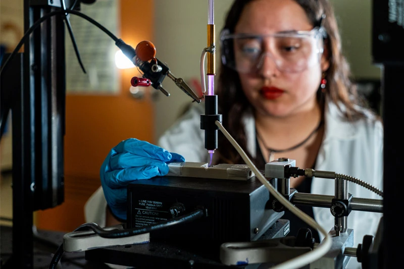 A scientist wearing safety glasses and blue gloves uses a device emitting a purple plasma beam onto a small metal plate.