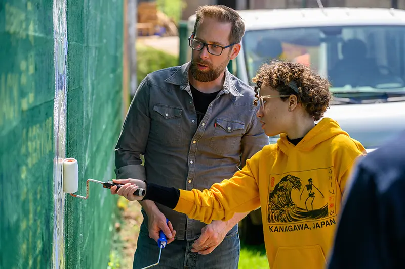 Carlson assisting a student paint a wall with a paint roller.