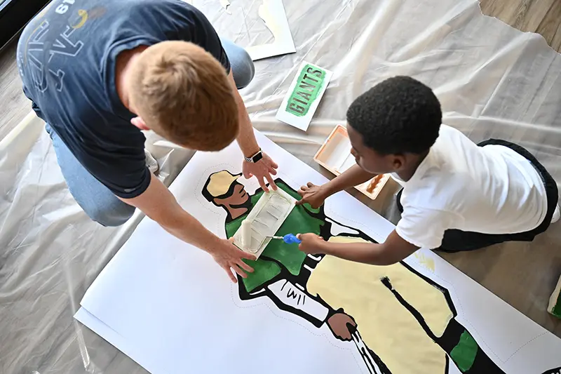 Notre Dame student kneels down to hold stencil in place for a younger student with the Boys and Girls Club of South Bend.