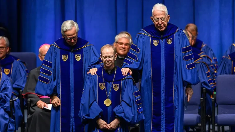 Fr. Dowd standing a few steps below Fr. Malloy and Fr. Jenkins as they place their hands on Fr. Dowd's shoulders during the blessing.