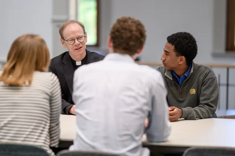 Fr. Bob Dowd sitting at a table talking to three students.