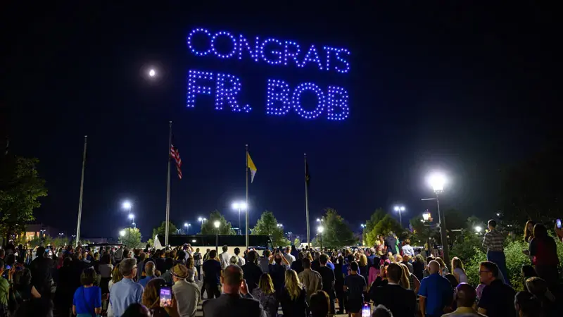 Over a dark night sky, drones equipped with blue LED lights spell out the words 'Congrats Fr. Bob' as a part of Inauguration festivities on campus.