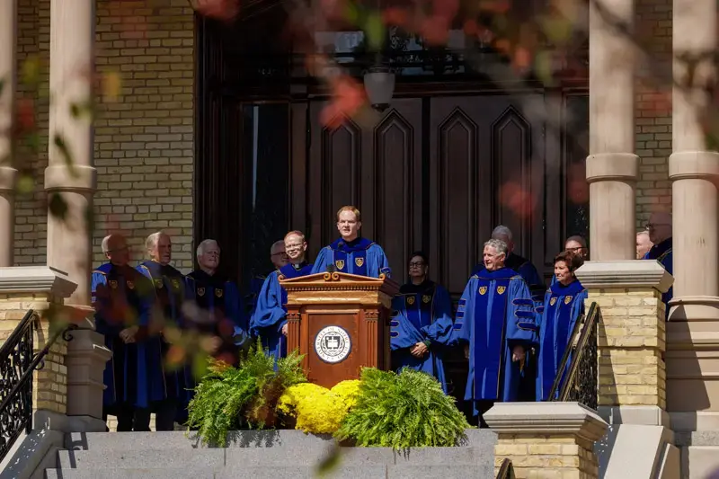 Standing a a podium, Rev. Greg Haake, C.S.C., dressed in academic regalia, stands at the steps of the administration building to read Father Sorin's letters.