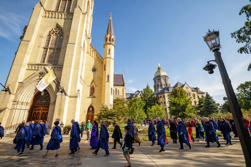 A line of people wearing academic regalia as they walk into the Basilica of the Sacred Heart on Notre Dame's campus.