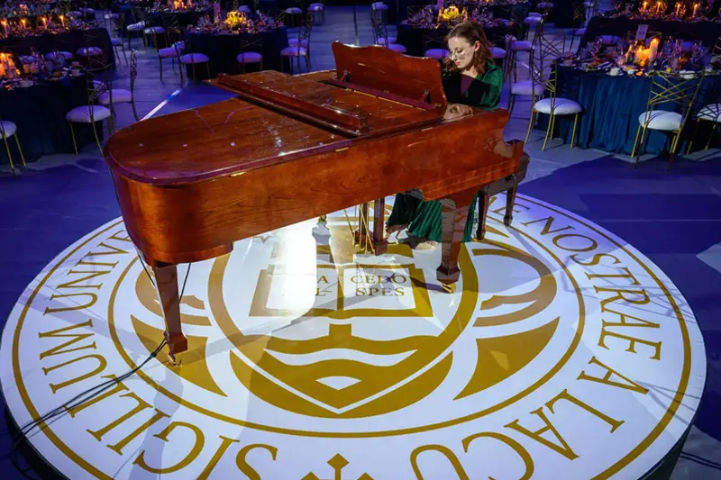 Miriam Rice sits at a piano on a stage in the middle of a room full of empty round tables.