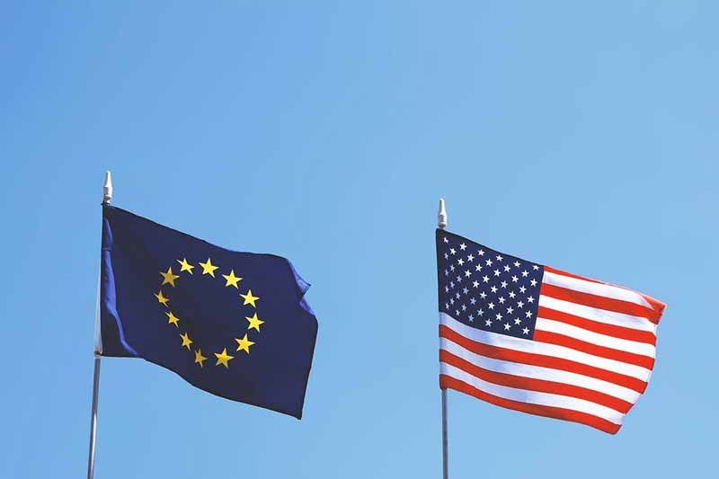The flag of Europe and the US flag fly side by side against a blue sky.