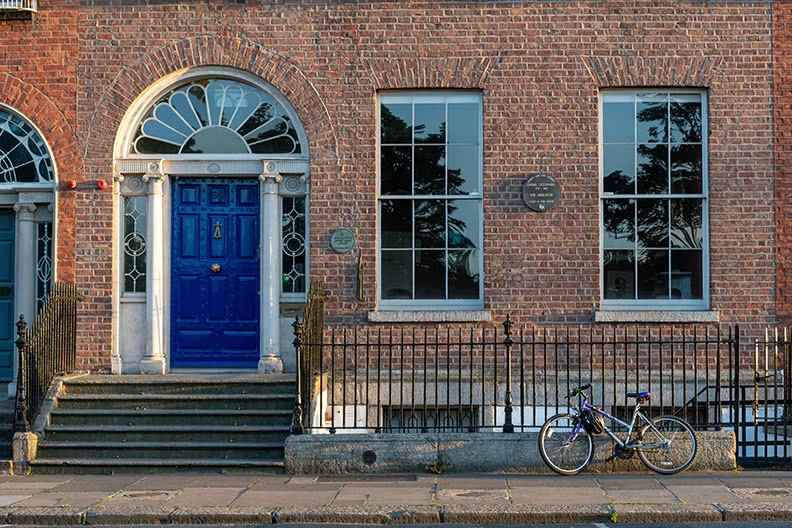 The blue door of the O'Connell House shown in early sunset with a bycicle leaning against the walk path fence.