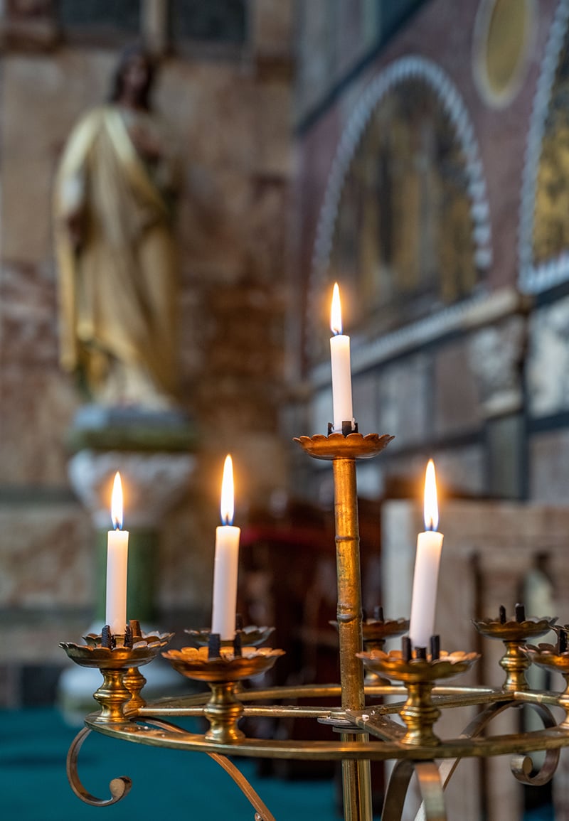 An in-focus candle holder in the foreground with a statue of Jesus on the cross out-of-focus in the background.