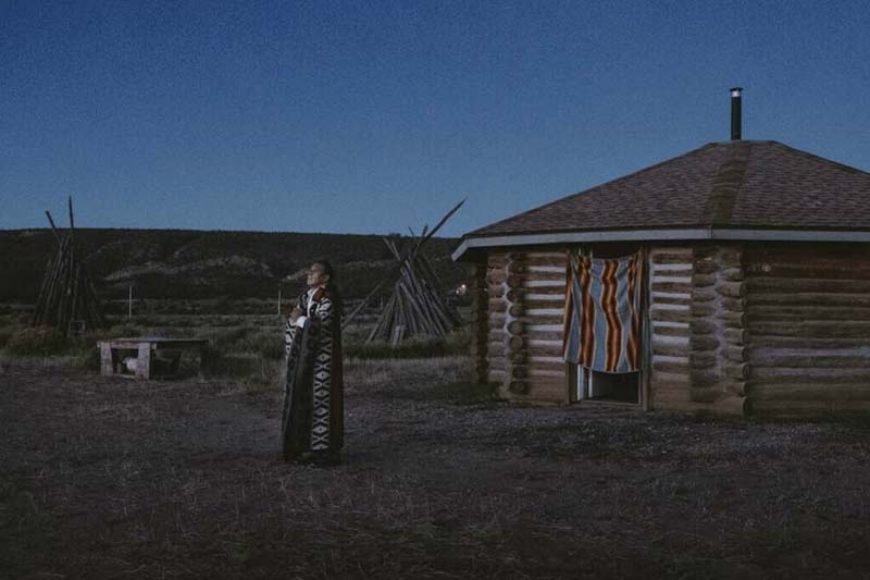 A Native American stands outside a log house looking up at the night sky.
