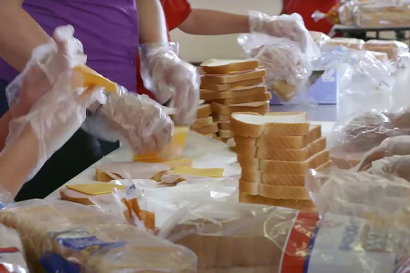 A table covered in white bread and lunch meats. People prepare sandwiches.