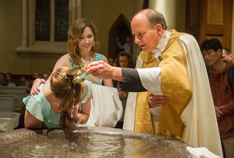 Fr. Rocca in gold vestments pours water over the head of a student.