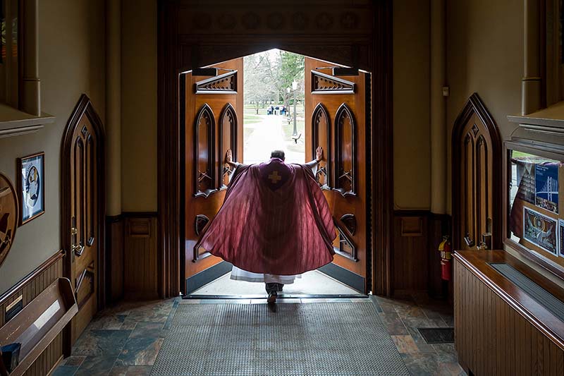 Fr. Rocca in a maroon vestments pushes open the doors of the Basilica to go outside.