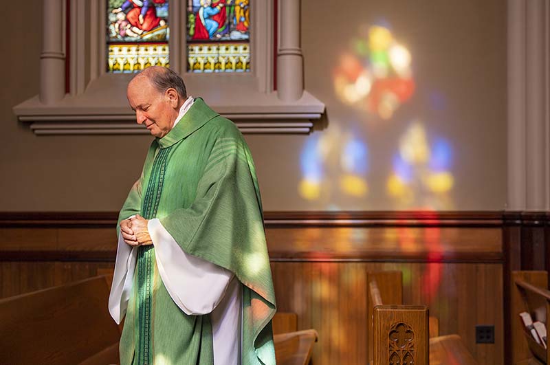 Fr. Rocca in green vestments gazes down in prayer, with colorful light reflecting from the stained glass windows on the wall.