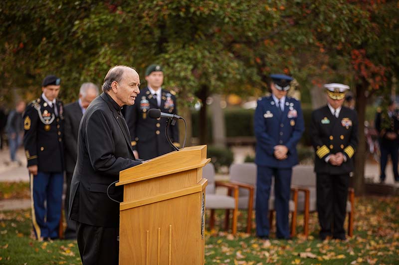 Father Rocca speaks at a podium with men in military dress in the background.