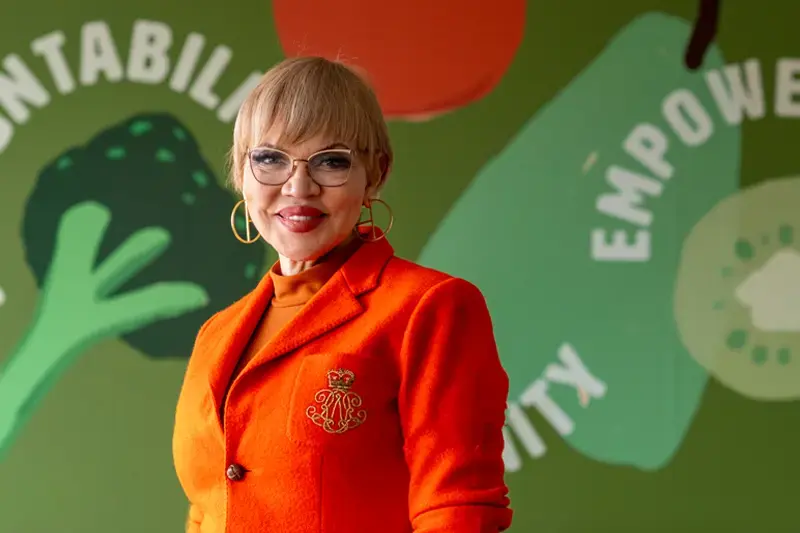 A woman with short blonde hair, glasses, and an orange blazer stands in front of a green wall with the words 'Values,' 'Accountability, 'Humility,' 'Empowerment, and 'Integrity' interspersed with images of produce.