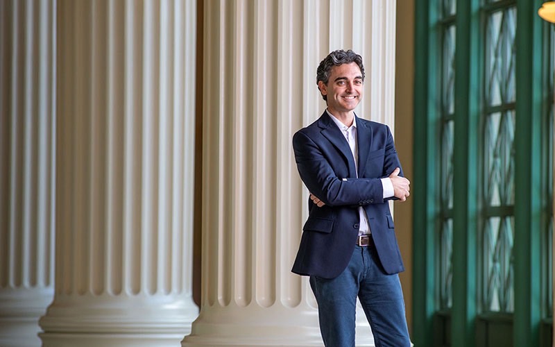 Professor Alessandro Pierattini stands in front of three large Roman columns in a hallway. On the right are green doors with triangular glass panes