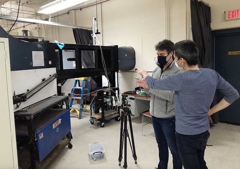 Aerospace engineers Gianluca Blois and Mitsugu Hasegawa stand together in front of the wind tunnel discussing wind tunnel analysis results.