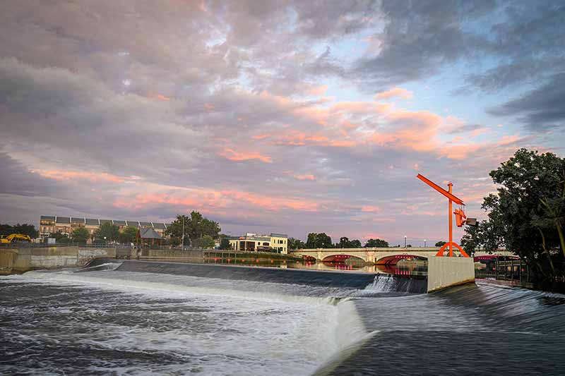 Pastel clouds in the sky, a bridge in the background, along a river and dam. An orange sculpture to the left of the river.