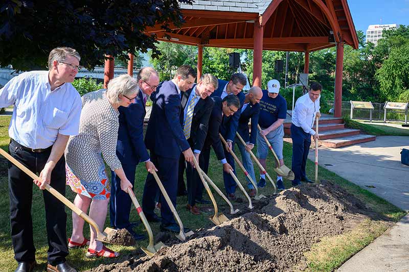 Ten people holding golden shovels dig into a small row of dirt.