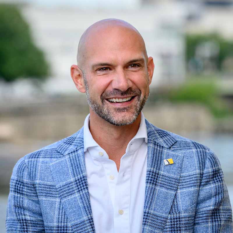 A headshot of a white, bald man wearing a white shirt and blue patterned suit jacket.