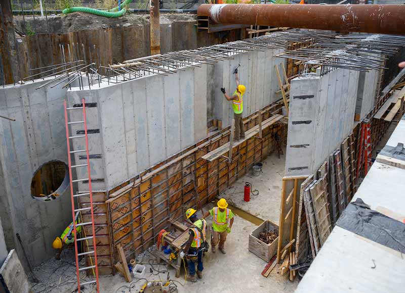 A narrow construction space, seen from above, surrounded by concrete walls, wooden panels, and metal wires. Four construction workers wearing protective gear work on site.