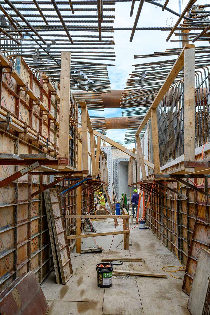 A narrow construction space surrounded by wooden boards and metal wires. Two construction workers wearing protective gear work on site.