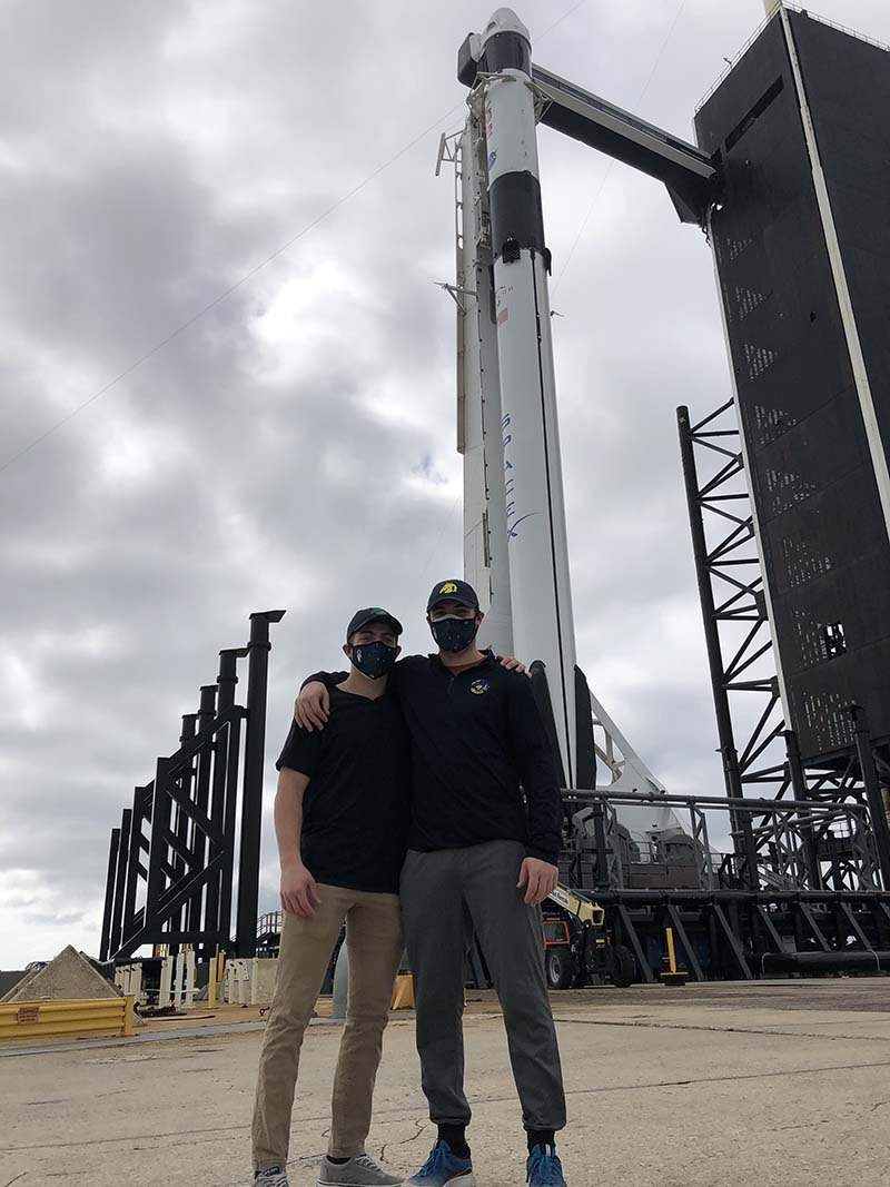 Two young men stand arm in arm in front of a rocket.