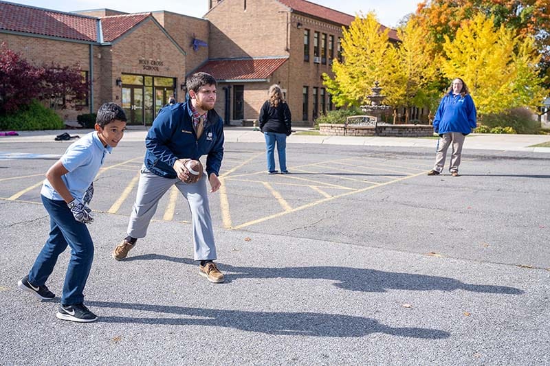 A male teacher plays football with students in a parking lot outside the school.