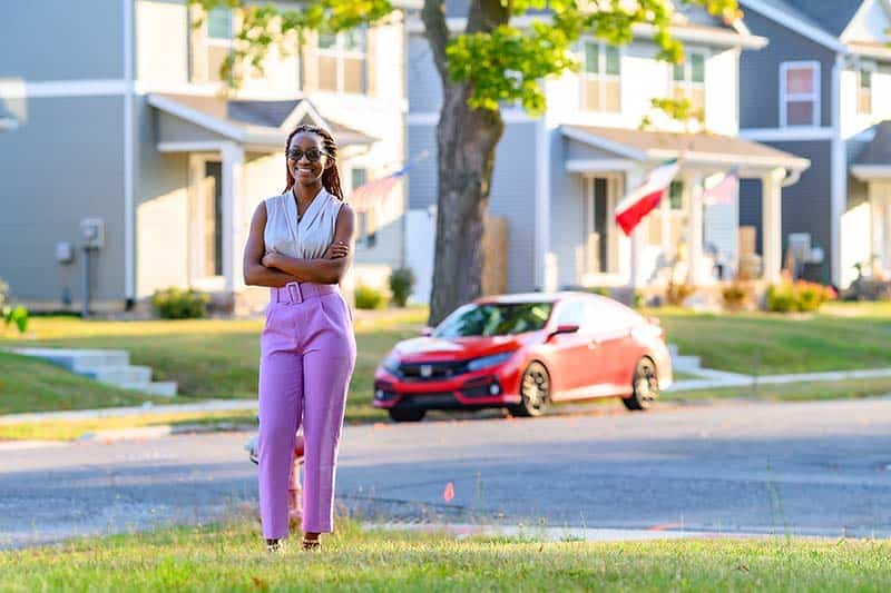 A female student, wearing glasses and light purple work pants, stands with her arms crossed at a street corner in front of newly constructed homes.