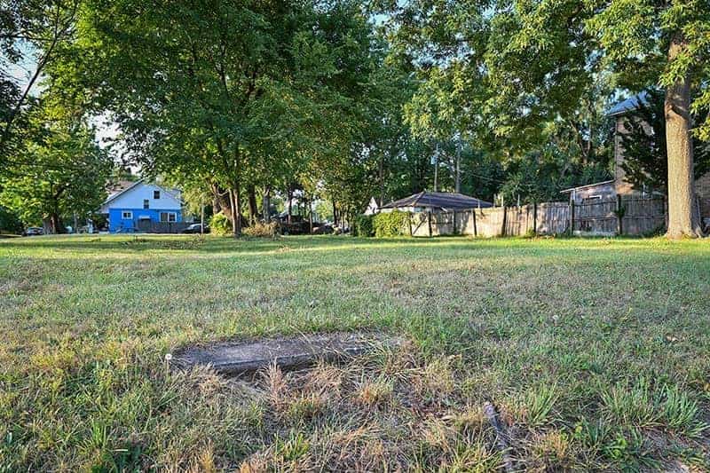 An empty lot with grass and a concrete step in the dirt. Backyards of other houses are in the distance.