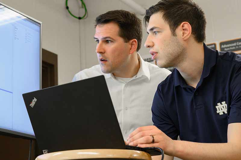 Two men working on a laptop look at a monitor to the left and evaluate data.