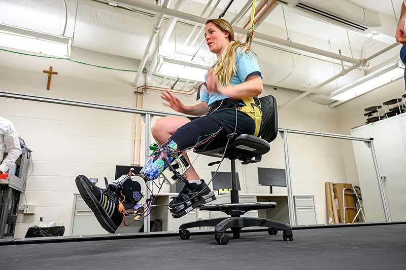 A woman with blonde hair sits in a rolling office chair and tests out a powered prosthetic lower-leg.
