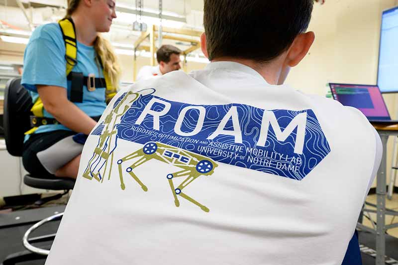 Detail of the back of a man wearing a ROAM t-shirt. In the background behind the man are two people sitting in the lab.