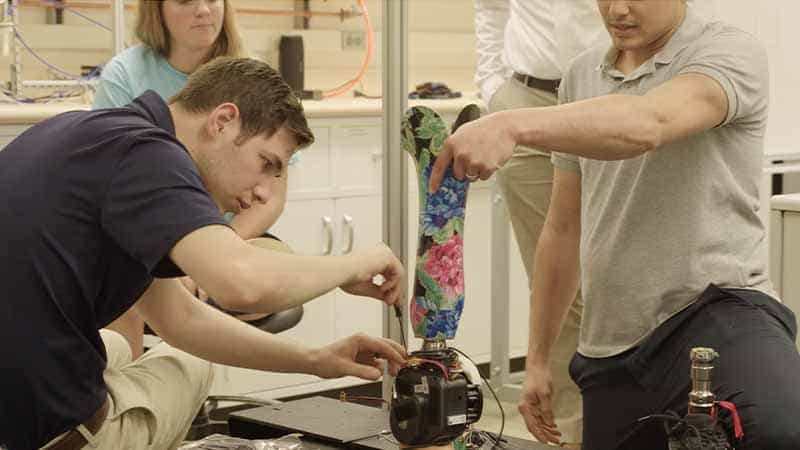A man uses a small screwdriver to work on a powered prosthetic lower-leg while another holds onto the top of the prosthetic leg.