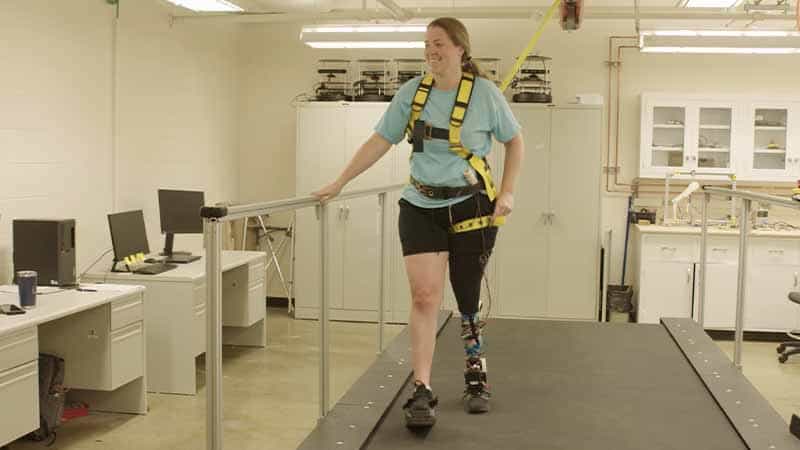 A woman wearing a bright yellow harness walks on a treadmill wearing a powered prosthetic lower-leg and smiles while holding onto the railing next to her.