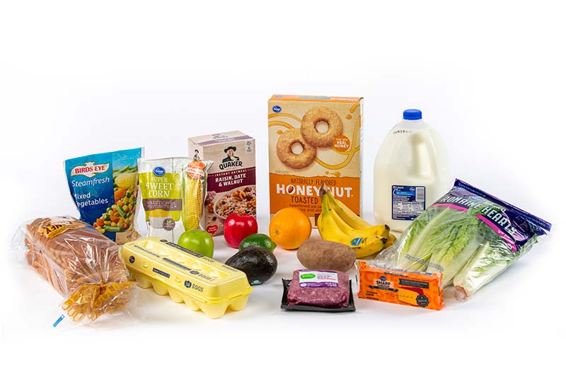 Fruits and vegatables, along with two frozen bags, eggs, bread, a box of cereal, milk, lettuce, a block of cheese, oatmeal, and ground beef on a white background.