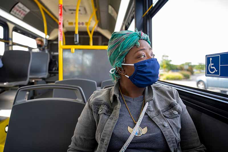 A woman wearing a headwrap and a mask sits on a bus and stares out the window.