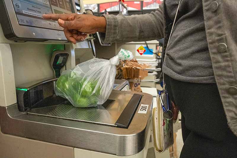 Green peppers wrapped in plastic sit on a self check-out scale while a hand pushes buttons on a screen.