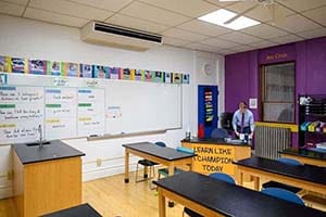 A teacher stands behind his desk in a classroom.