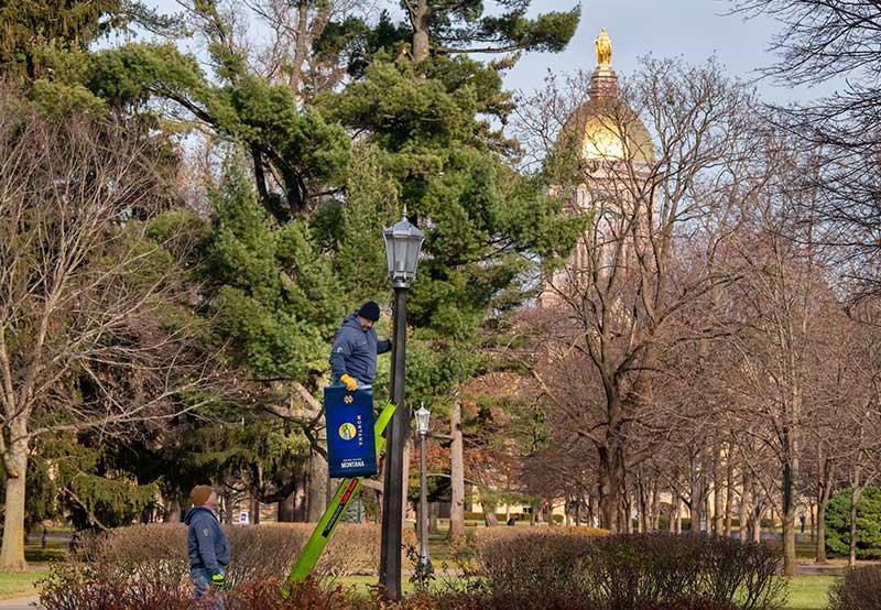 Two men installs a banner representing Montana's flag on a lamp post. The Golden Dome is in the background, surrounded by pine trees and bare trees.