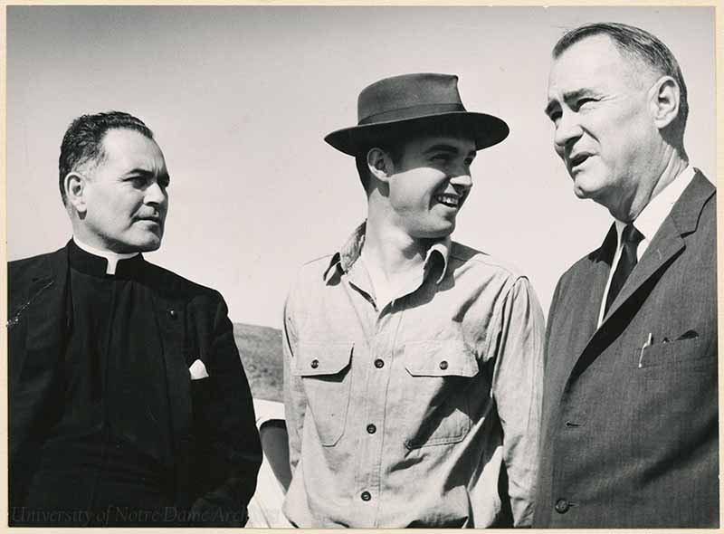 A black and white, archival photo of three men. Rev. Theodore M. Hesburgh, C.S.C., wearing a priest collared shirt, looking to the right at Edward Tisch, wearing a button-up long sleeve shirt and fedora hat, and Walter Langford, wearing a suit and tie.