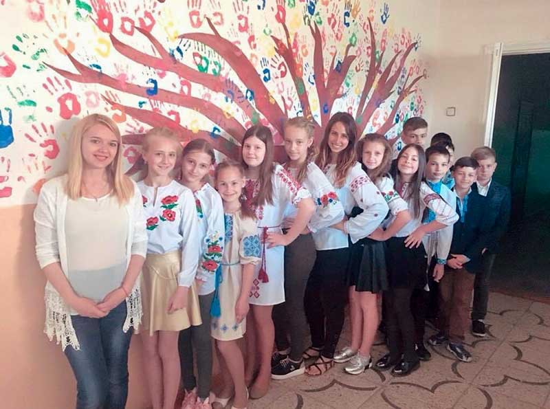 A woman, Mathilda Nassar, poses for a photo with her students in front of a painted tree on the wall.