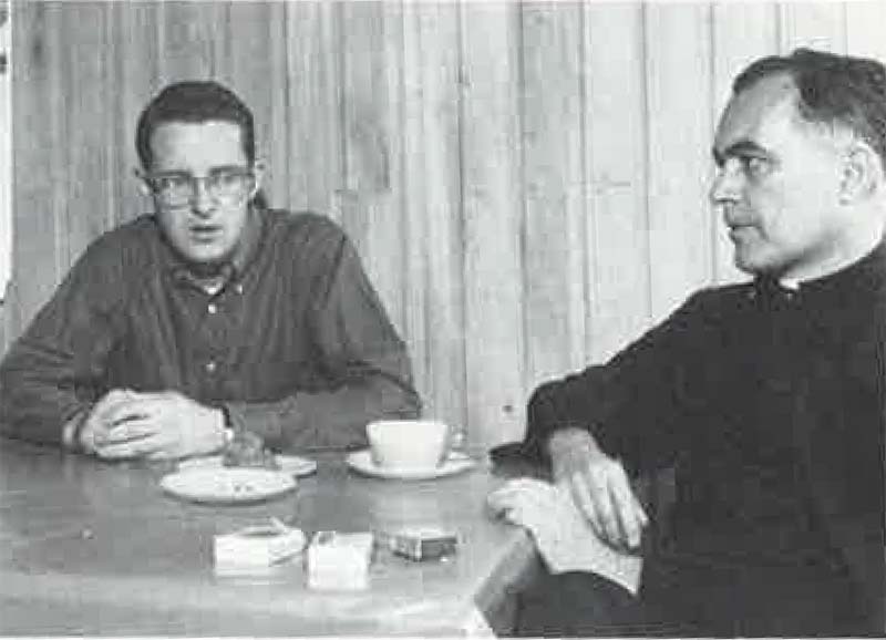 A black and white archival photo of Tom Scanlon speaking with Rev. Theodore M. Hesburgh C.S.C. sitting at a table having coffee or tea, against a wood panel wall.