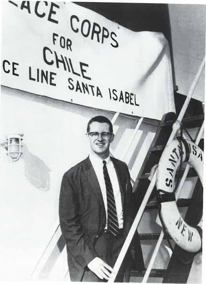 A black and white archival photo of Tom Scanlon, wearing a suit, tie, and glasses, posing on a ship holding onto a stair railing. Behind him is a Peace Corps for Chile sign.