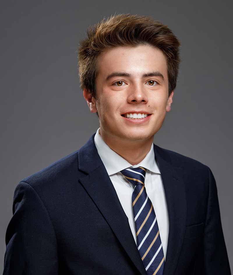 A white male student, wearing a navy suit, white shirt, and striped tie, poses for a headshot.