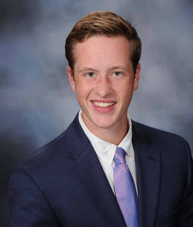 A white male student, wearing a navy suit, white shirt, and purple tie, poses for a headshot.