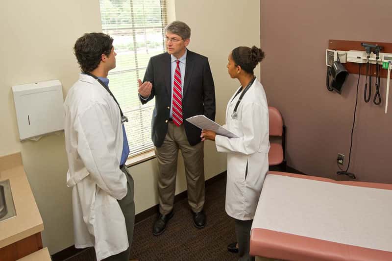 A teacher converses with two students in lab coats in a doctor's office.