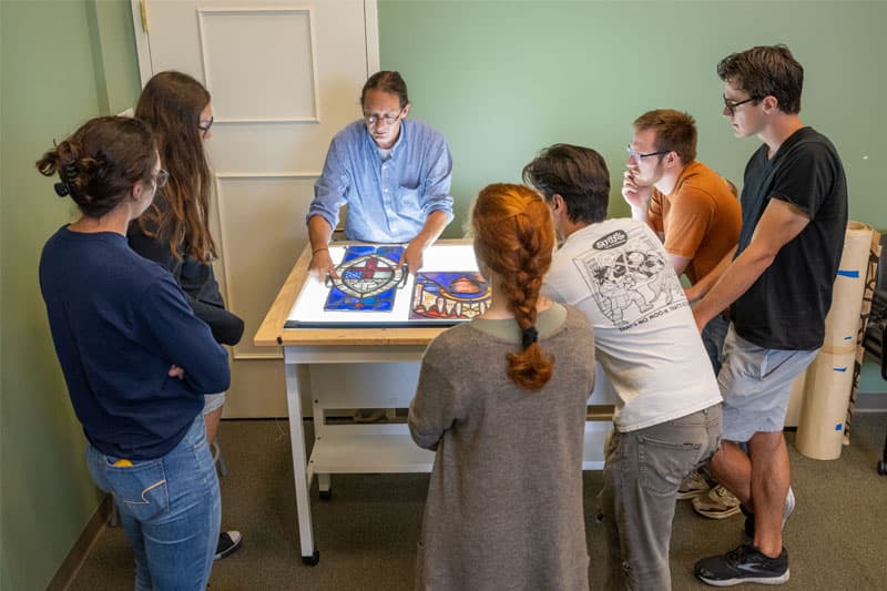 Professor Stephen Hartley stands over a light table illuminating a stained glass drawing while surrounded by students.